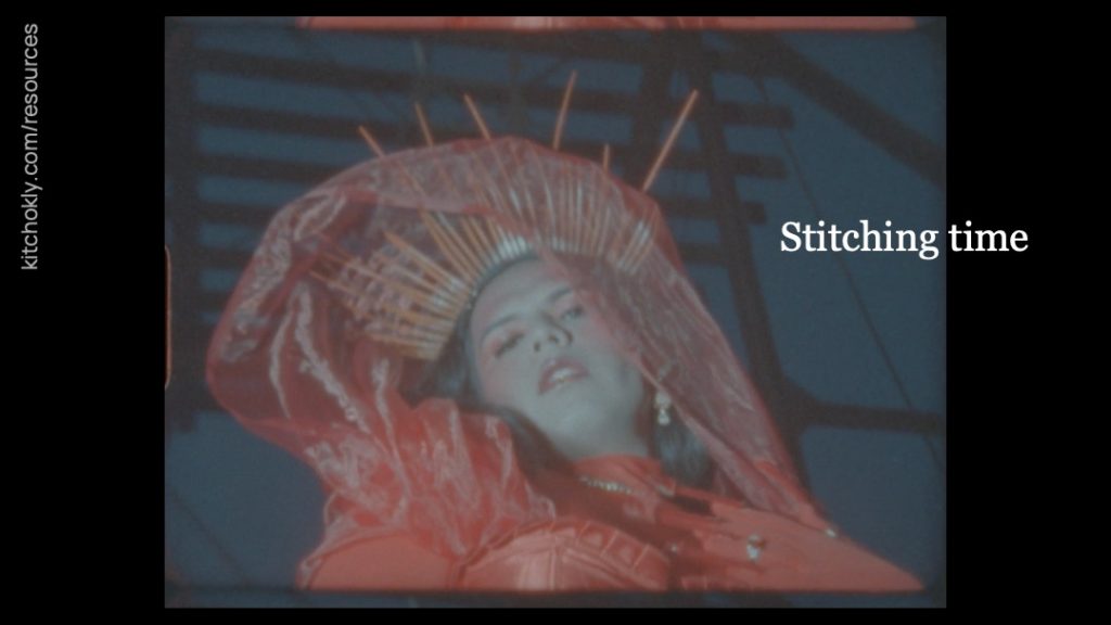 The figure reappears in their red outfit, this time looking down at the camera. Shimmering, translucent fabric is tangled in their diadem. In the background appears to be a metal structure. Where "Cutting words" were before on slides 8 and 9 now reads "Stitching time".